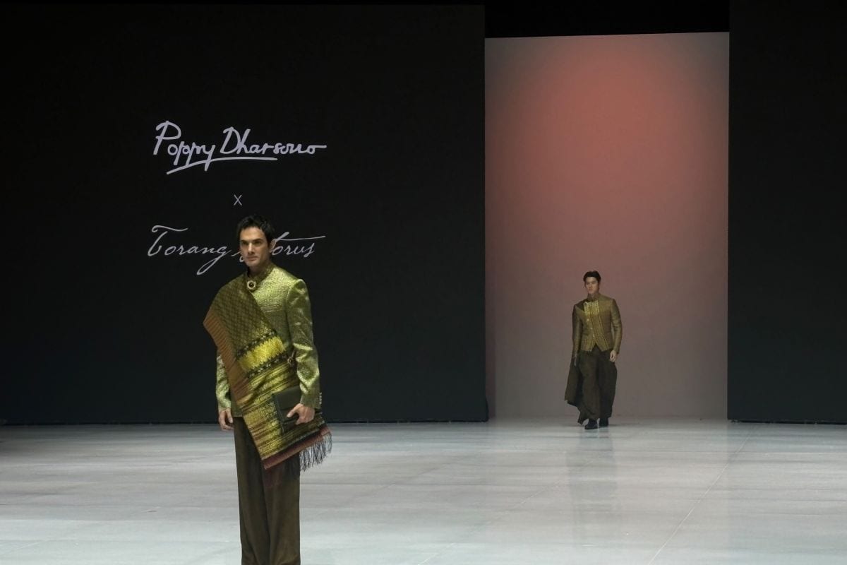 A model wearing a woven ulos shawl over a gold jacket walks the runway in front of a Poppy Dharsono x Torang Sitorus backdrop