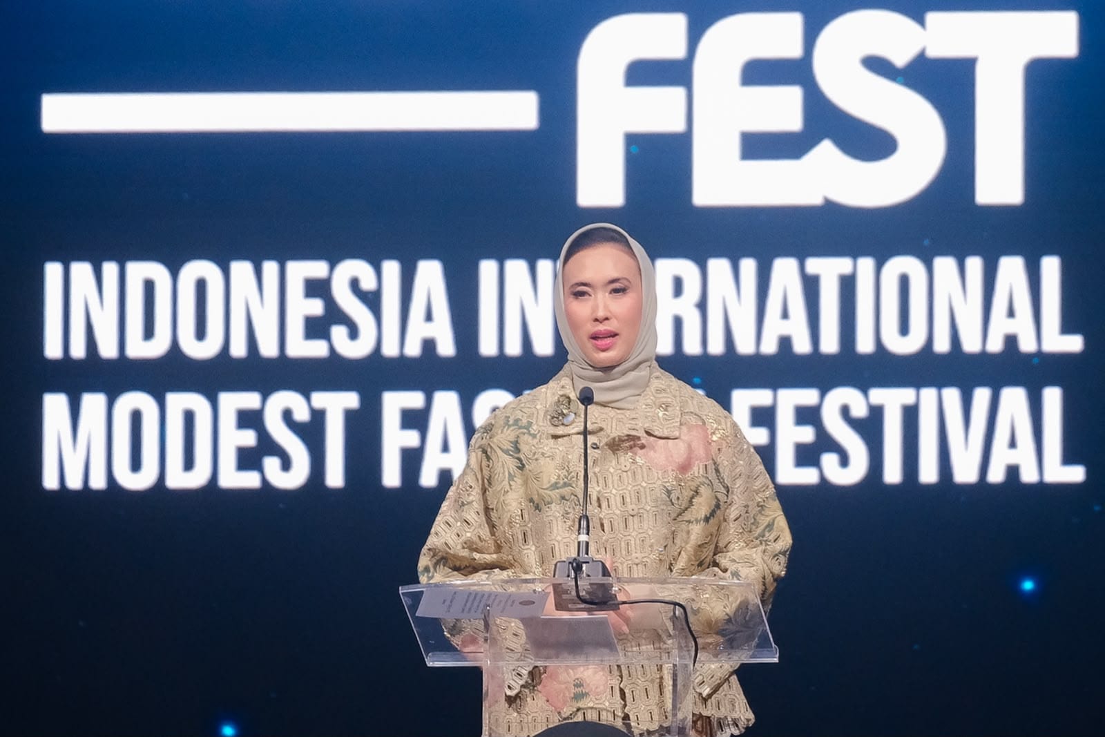 A speaker in a batik-inspired outfit and hijab gives opening remarks in front of the Indonesia International Modest Fashion Festival screen