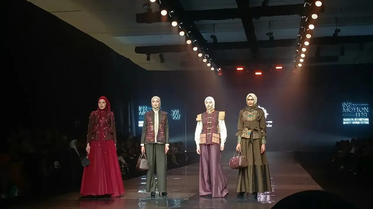 Four models in hijabs, woven-panel vests, wide trousers and a red gown stand at the end of the runway beneath IN2MOTIONFEST screens