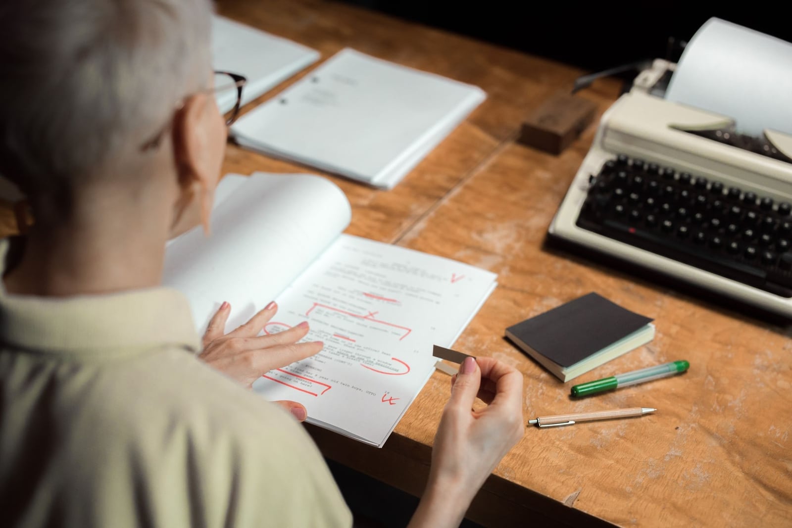 Seen from behind, a reader marks printed pages with red-pen corrections at a wooden desk beside a cream typewriter and notebook