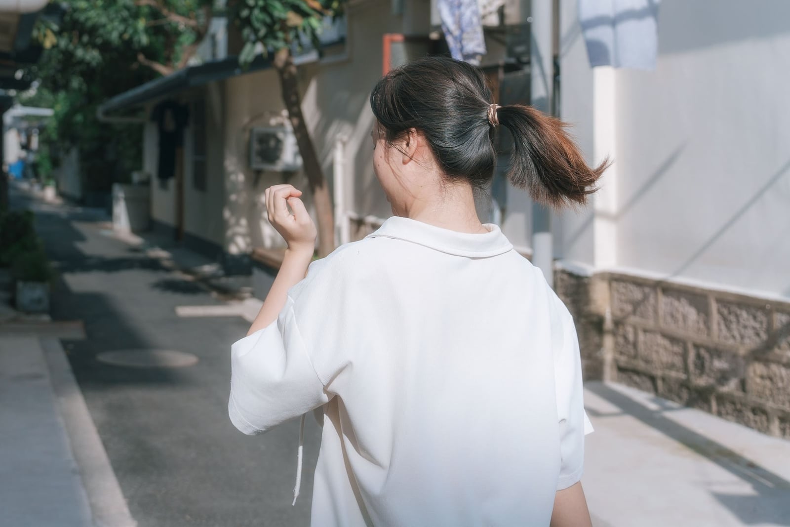 Seen from behind, a woman in a loose white shirt with a swinging ponytail walks down a quiet, sunlit residential lane