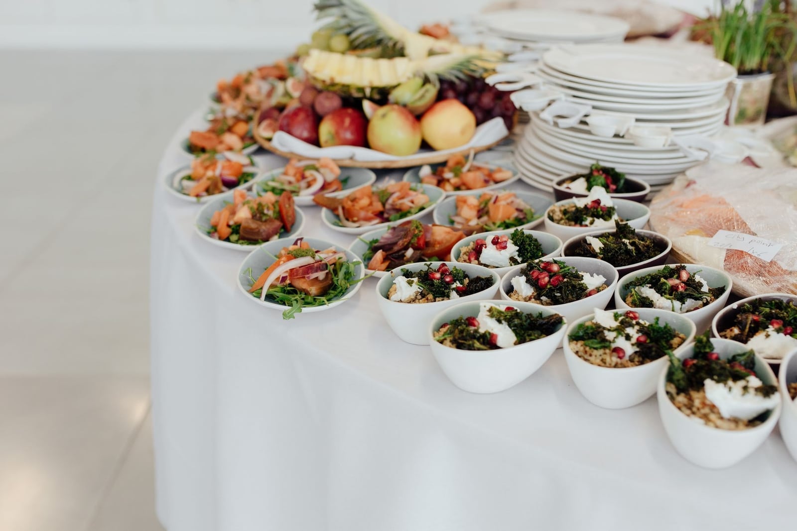 A white-clothed catering table set with small bowls of grain salads and greens, a platter of fresh fruit and stacks of white plates
