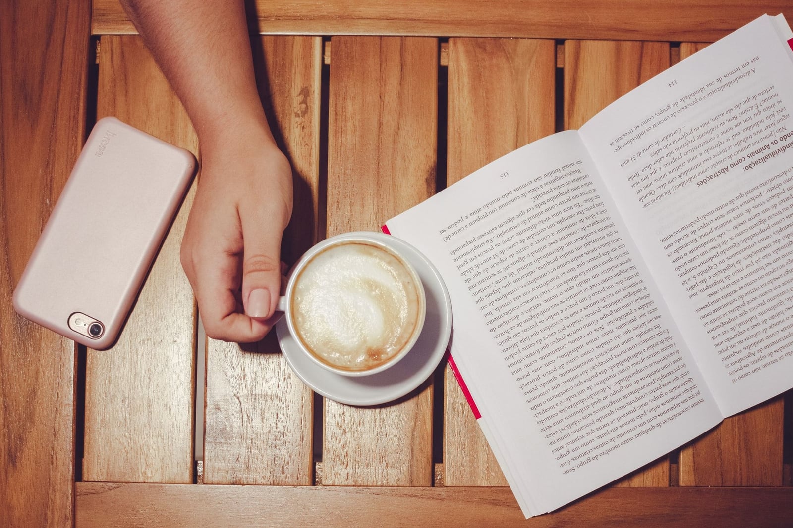 Overhead view of a phone lying face-down on a slatted wooden table beside a hand holding a cappuccino and an open paperback