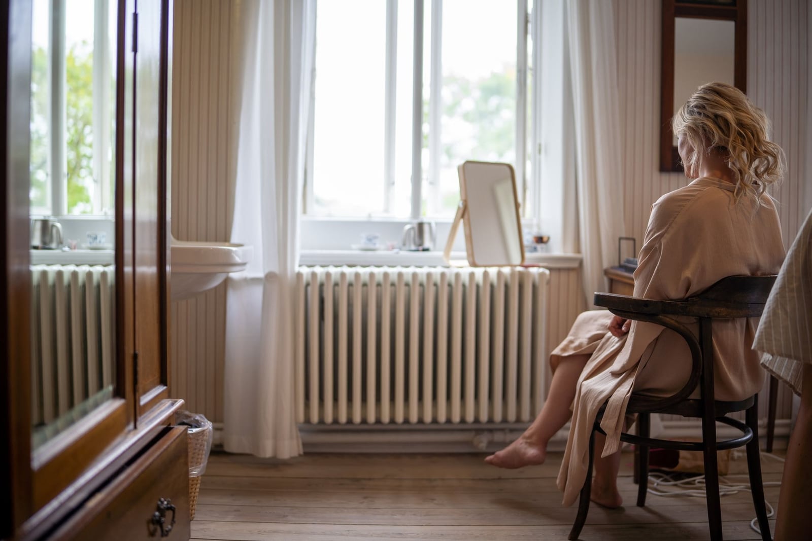 Seen from behind, a woman in a silky robe sits barefoot on a bentwood chair in a quiet, sunlit room with tall windows and an old radiator