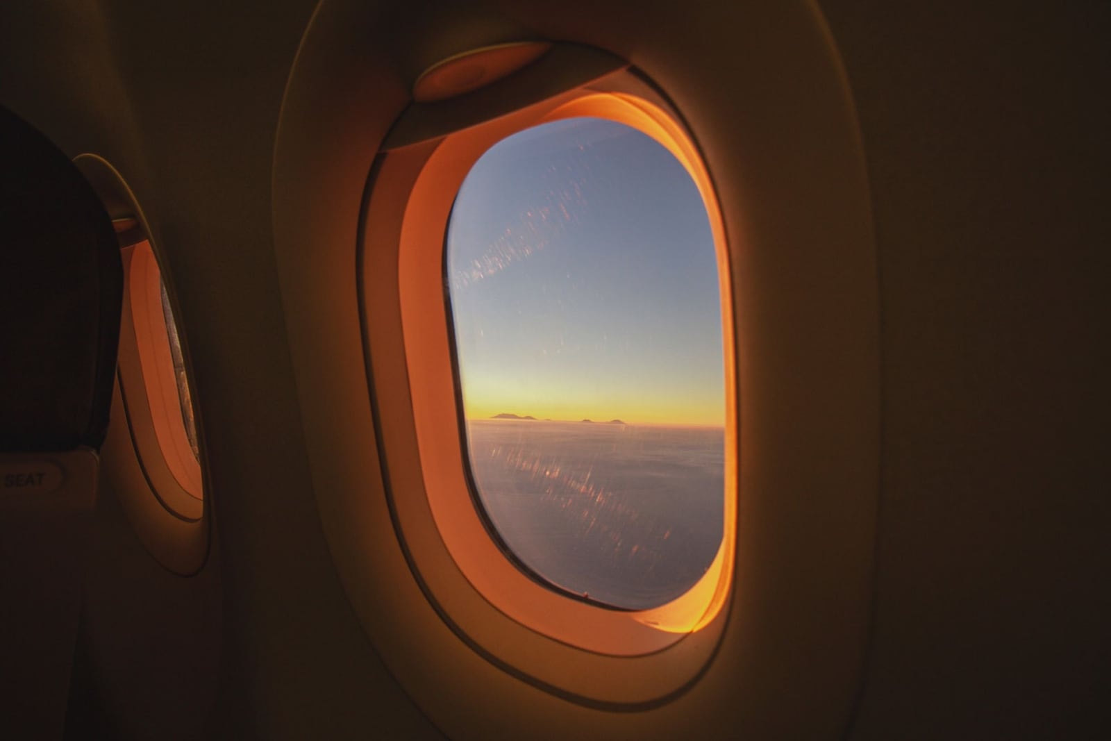 An aeroplane window glowing orange at sunrise above a sea of clouds, the darkened cabin wall curving around it